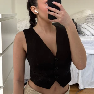Svart väst med knappar - Snygg svart väst med knappar framtill. Perfekt för att lägga till en stilren touch till din outfit. Västen har en v-ringad design och en tight passform. Från Nelly