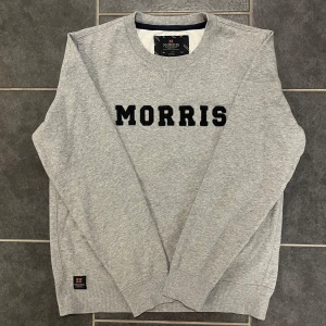 Grå sweatshirt från Morris - Säljer en stilren grå sweatshirt från Morris med svart text på bröstet. Tröjan är tillverkad i en mjuk bomullsblandning och har ribbade muddar vid ärmslut och nederkant. Perfekt för en avslappnad look.