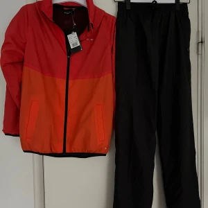 Röd och orange vindjacka och byxor  från SOC - vindjacka och byxor från SOC i orange och svart  Jackan har en dragkedja framtill och en hög krage med meshfoder. Perfekt för utomhusaktiviteter när det blåser. Jackan har långa ärmar och elastisk nederkant för bättre passform. Byxorna dragsko vid benavslut och resår med dragsko. Helt nytt! Storlek 158-164