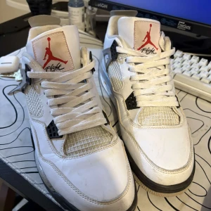 Vita Nike Air Jordan 4 - Säljer ett par vita Nike Air sneakers med svarta detaljer och röd logga. Skorna har snörning och en klassisk design med nätpaneler. Perfekta för en sportig look.