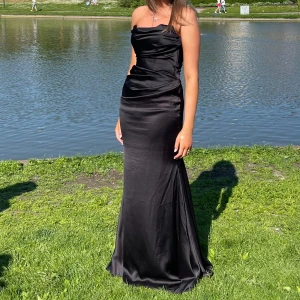 Svart långklänning - Elegant svart långklänning i satin med axelbandslös design. Klänningen har en smickrande passform och en vacker drapering framtill. Perfekt för en formell tillställning.