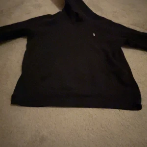 Svart hoodie från Ralph Lauren - Säljer en stilren svart hoodie från Ralph Lauren med det klassiska polospelar-emblemet på bröstet. Perfekt för en avslappnad och bekväm stil. Hoodien har långa ärmar och en mjuk huva.