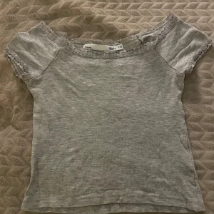Grå topp med spetsdetaljer - Säljer en söt grå topp med spetsdetaljer vid halsringningen och ärmsluten. Sparsamt använd och liten i storlek!