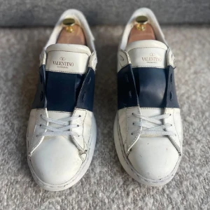 Valentino Open - Snygga Valentino Garavani sneakers i vitt med en bred blå rem över snörningen. Skorna har en klassisk design med rund tå och är tillverkade i skinn. Perfekta för en stilren look. skicket är rätt så bra men det förekommer defekter. 