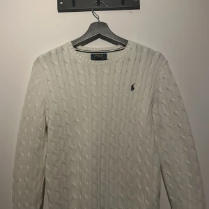 Vit stickad tröja från Polo Ralph Lauren - Säljer en stilren vit stickad tröja från Polo Ralph Lauren med kabelstickat mönster. Tröjan har långa ärmar och en rund halsringning. Den ikoniska loggan är broderad på bröstet. Perfekt för en klassisk och tidlös look.