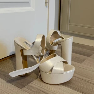 Vita platåpumps med klack - Säljer ett par snygga vita platåpumps med hög klack. De har en stilren design med breda remmar och ett spänne runt ankeln för extra stöd. Perfekta för festliga tillfällen när du vill sticka ut!