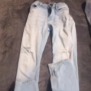 Ljusblå jeans med slitningar - Säljer ett par ljusblå jeans från SMOL Denim. De har en cool, sliten look med hål på framsidan. Perfekta för en avslappnad stil. De är i storlek 29/32 och gjorda i Bangladesh. Och har inte använt så mycket 