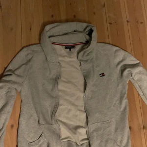 Grå hoodie från Tommy Hilfiger - Säljer en snygg och bekväm grå hoodie från Tommy Hilfiger. Den har en dragkedja framtill och en klassisk logga på bröstet samt en stor logga på ryggen. Perfekt för en avslappnad stil och passar till de flesta tillfällen. 10/10 skick