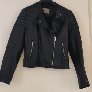 Svart skinnjacka  - Svart fejk skinnjacka/läderjacka med detaljer runt axlarna. I fint skick, det enda anmärkningen är att jag vikt ärmarna lite så tyget töjts ut lite där. Se bilder. Annars hel och fin! Passar en M/L