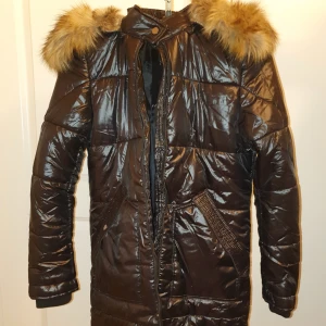 Brun dunjacka med pälsdetaljer - Säljer en snygg brun dunjacka med pälsdetaljer på huvan. Jackan är lång och perfekt för kalla vinterdagar. Den har en glansig finish och långa ärmar. Stängs med dragkedja och knappar för extra värme. Supermysig och stilren!