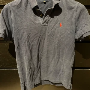 Blå pikétröja från Ralph Lauren - Säljer en klassisk Blå pikétröja från Ralph Lauren med korta ärmar och en broderad röd logotyp på bröstet. Perfekt för en stilren och avslappnad look. Varan är inköpt från Kidsbrandstore. Storleken är i barnstorlek XL, 18-20 år och motsvarar enligt mig S-M i herr storlek. 