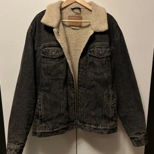 Svart jeansjacka med teddyfoder från Pull&Bear - Säljer en svart jeansjacka från Pull&Bear med teddyfoder. Jackan har en klassisk krage och knappar framtill, samt knappar i änden av båda armar för att kunna öppna ärmen bitvis vid underarmen. Bekväm och värmer rätt bra vid kallare väder.