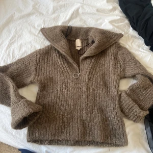 Brun stickad tröja med dragkedja - Mysig brun stickad tröja med en stor krage och dragkedja framtill. Perfekt för kyliga dagar och ger en avslappnad look.