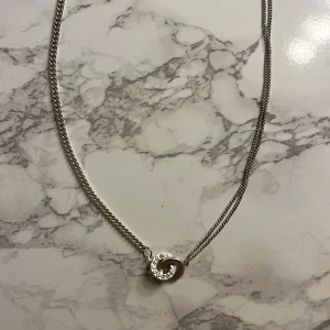 Silverhalsband med cirkelberlock - Elegant silverhalsband med en dubbel cirkelberlock, varav en är dekorerad med små stenar. Kedjan är tunn och smidig, perfekt för att ge en subtil glans. Märket är ingraverat på en liten rund platta vid låset.
