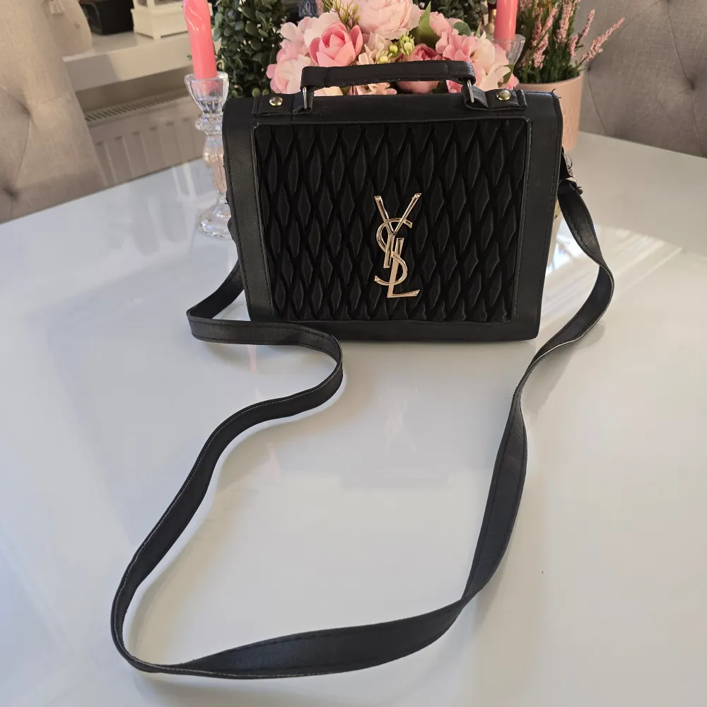 Snygg svart axelväska med quiltad design och guldfärgad logotyp på framsidan. Väskan har en justerbar axelrem och ett handtag för enkel bärning. Perfekt för att ge en elegant touch till din outfit. Inte äkta!. Laukut & Käsilaukut.