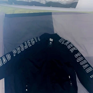 Svart jacka från Black Squad - Snygg svart jacka från Black Squad med textdetaljer på ärmarna. Jackan har dragkedja framtill och praktiska fickor med dragkedjor. Perfekt för en cool och avslappnad stil.