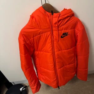 Orange pufferjacka från Nike - Säljer en cool orange pufferjacka från Nike med huva och dragkedja. Jackan har en sportig look med Nike-logga på bröstet. Perfekt för kyliga dagar.