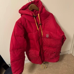 Röd dunjacka från Fjällräven - Snygg röd dunjacka från Fjällräven med huva och gula dragskor. Jackan har praktiska fickor med knappar och en dragkedja framtill. Perfekt för kyliga dagar.