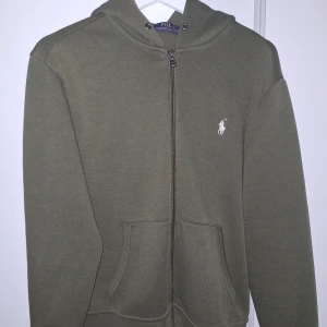 hoodie från Ralph Lauren - Snygg olivgrön hoodie från Ralph Lauren med dragkedja och klassisk logga på bröstet. Perfekt för en avslappnad stil. Den har en luva och fickor framtill.