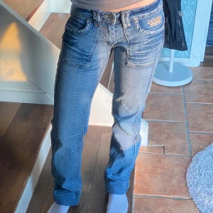 Blå jeans med coola detaljer - Snygga unika blå utvättade jeans med broderad detalj på fickorna. De har en klassisk femficksdesign och en knappgylf. Jeansen är straight/ bootcut med låg midja och dom är insydda i midjan (inget som syns) men man skulle kunna lösa upp och göra dem större i midjan (som dem var från början). Storleken står ej men jag skulle anse att dem är ca xs/s (ganska stretchiga också). Midjemått rakt över: 37cm. Innerbenslängd: ca 71 cm. Ytterbenslängd: ca 100cm. Fråga om funderingar finns❣️