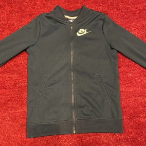 Blå / mörkturkos kofta från Nike - Säljer en kofta från Nike med dragkedja och långärmad design. Jackan har en klassisk Nike-logga på bröstet och praktiska fickor framtill. Perfekt för en sportig look!