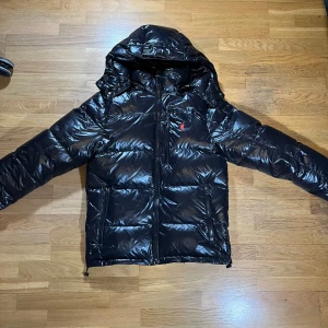 Svart dunjacka från Polo Ralph Lauren - M men passar även S. Är en snygg svart dunjacka från Polo Ralph Lauren med huva och dragkedja. Perfekt för kyliga dagar med sin pufferdesign som håller dig varm och stilren. Är en Replika.