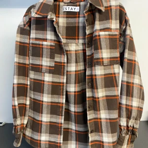 Brunrutig overshirt från Stay - Snygg brunrutig overshirt från Stay med långa ärmar och knappar framtill. Skjortan har en klassisk krage och två bröstfickor. Perfekt för lager-på-lager-stil under kyligare dagar. Nypris: 600kr, knappt använd