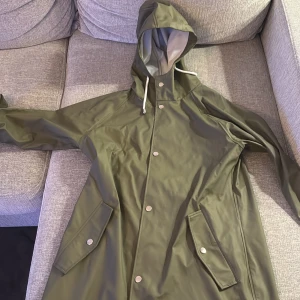 Grön regnjacka med huva - Snygg grön regnjacka från 157 med huva och knappar. Perfekt för regniga dagar. Jackan har praktiska fickor och dragsko vid huvan för extra skydd.