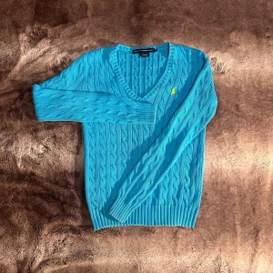 Ralph Lauren - Säljer en snygg blå stickad tröja från Ralph Lauren med v-ringning och kabelstickat mönster. Tröjan har långa ärmar och en liten gul logga på bröstet. Perfekt för en stilren look!