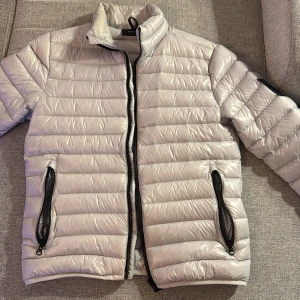 Grå pufferjacka från Peak Performance - Snygg och stilren grå pufferjacka från Peak Performance. Jackan har en dragkedja framtill och praktiska fickor med dragkedjor. Perfekt för kyligare dagar med sin quiltade design.