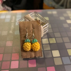 Ananasformade örhängen - Söta örhängen i form av ananaser med gula frukter och gröna blad. Perfekta för att ge en tropisk touch till din outfit. De hänger från silverfärgade krokar och är lätta att bära.