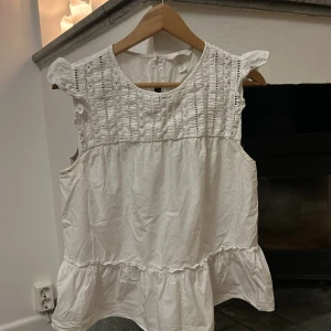 Vit blus från H&M - Säljer en söt vit blus från H&M med spetsdetaljer och volang nedtill. Blusen är ärmlös och har en rund halsringning. Perfekt för sommarens varma dagar då den är väldigt luftig och skön!❤️Nypris 300 kr