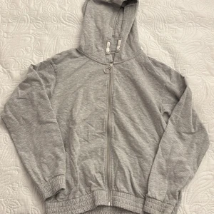 Grå hoodie från Lindex - Säljer en grå hoodie från Lindex i storlek 170. Den har dragkedja framtill och en praktisk huva. Tillverkad i en mjuk blandning av bomull och viskos med lite elastan för stretch.