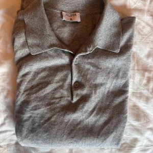 Grå långärmad polotröja från Filippa K - Säljer en stilren grå polotröja från Filippa K i mjukt ullmaterial. Tröjan har en klassisk krage med knappar framtill. Perfekt för en elegant och avslappnad look.