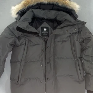 Svart dunjacka med pälsdetalj från Canada Goose - Säljer en stilren svart dunjacka från Canada Goose med avtagbar huva och pälsdetalj. Jackan har dragkedja och knappar framtill samt en broderad logotyp på ärmen. Perfekt för kalla vinterdagar.