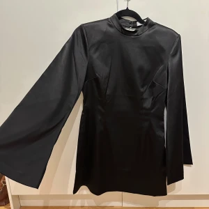 Svart klänning med öppen rygg - Elegant svart klänning i satin med långärmad design och en stilren hög hals. Klänningen har en iögonfallande öppen ryggdetalj som ger en modern touch. Perfekt för en chic look. Köpt för 2999kr & endast använd en gång, så den är i nyskick!💕