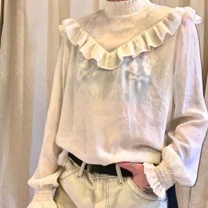 Genomskinlig blus - Supersöt gräddvit blus med volang och hög krage. Ganska genomskinlig så rekommenderar att ha något under. Passar XS-M. Rak boxig modell. 