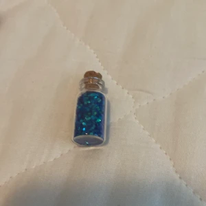 Halsband med blått glitter i flaska - Ett halsband med en liten flaska fylld med blått glitter. Flaskan har en kork och hänger i en kedja.