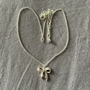 Silverfärgat halsband med rosett - Ett elegant silverfärgat halsband med en detaljrik rosett som hänge. Halsbandet har en fin kedja och rosetten är dekorerad med små pärlor.