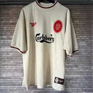 En beige fotbollströja från Liverpool F.C. tillverkad av Reebok. Tröjan har korta ärmar med röda och vita detaljer på kragen och ärmsluten. På framsidan finns Liverpool F.C:s emblem och Carlsberg-logotypen.