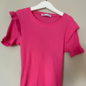 Rosa tröja med volanger på axlarna  - Rosa T-shirt från Zara med volanger på axlarna, köpt för ett tag sen men andats använd ca 3-5 ggr. Bra skick. Köpt för ca 200-250kr tror jag. Stretchigt tyg. Storlek S men passar XS också. Pris kan diskuteras.