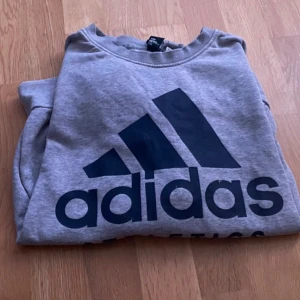 Grå sweatshirt från Adidas - En bekväm grå sweatshirt från Adidas med deras logotyp och texten 'ATHLETICS' tryckt på framsidan. Tröjan har långa ärmar och ribbstickade muddar vid ärmslut och nederkant.