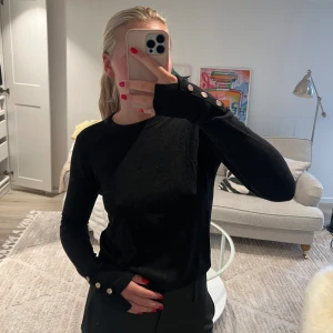 Svart tröja med gulddetaljer - En svart finstickad tröja med guldiga knappar vid armen.