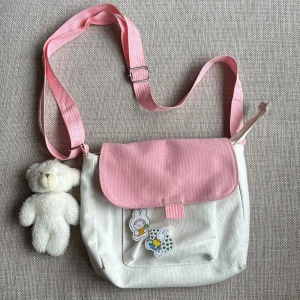 Väska  - Söt väska i vitt och rosa med dekaler och gosedjur hängande på sidan. Fint skick, aldrig använd. Två fack, det stora stängs med dragkedja (ett litet fack på insidan som också stängs med dragkedja) samt ett mindre med knapp på utsidan. Ca 20x22 cm