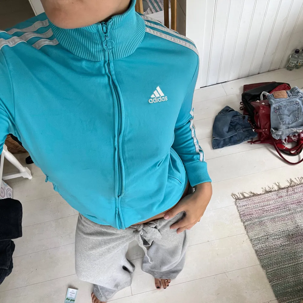 En turkos zipper från Adidas med vita detaljer och dragkedja. Jackan har långa ärmar och ribbstickade muddar vid ärmslut och nederkant. Den har även Adidas-logotypen på bröstet och de klassiska tre ränderna längs ärmarna. barnstorlek men passar S/M. Tröjor & Koftor.