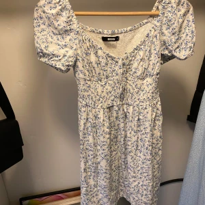 Blommig klänning - Vit klänning med blå blommor. Ballongärm och knytning fram. Har resår runt magen och rygg. Använd enstaka gång så i fint skick men några trädar som sticker ut