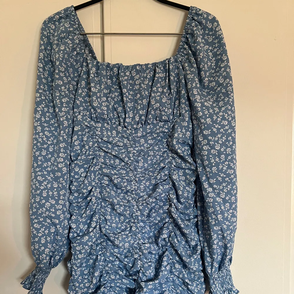 En blå klänning med vita små blommor på. Sitter smickrande på kroppen med hjälp av stretchen och modellen. Kan användas både som på bild men ocksp off shoulder om man drar ner armarna. Aldrig använd och väldigt fint skick! . Mekot.