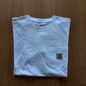 Vit T-shirt  - T-shirt från Carhartt. Något färgad i halsringen, se bild. Finns även en i svart, se min profil.   Rensar ur min garderob från en hel del oanvända kläder🌼 Om du ser något mer i min profil som du gillar så skickar jag med det till dig också!😊 