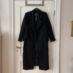 Kappa från Zara - Svart lång kappa från Zara. Storlek XS, men passar XS-M. På mig som är 160cm, är den ner till ungefär till nedanför vaden. Axelvaddar. Stängs med knappar. Slits baktill. Använd men i fint skick. Bara att fråga om det är något du undrar!