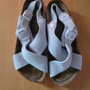 Vita sandaler från Birkenstock - Ett par vita sandaler från Birkenstock med korsade remmar och justerbara spännen. Sandalerna har en bekväm fotbädd och en vit sula med mönster för bättre grepp.